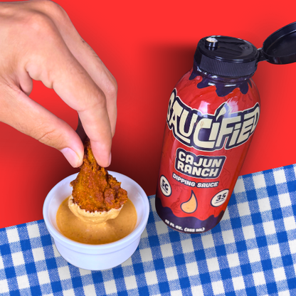 Hand dipping food into a bowl of sauce with a bottle of 'Cauciified Cajun Ranch' dipping sauce on a red and blue checkered tablecloth.