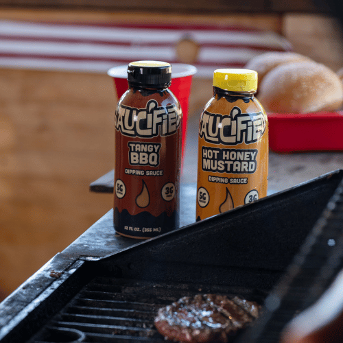 Two bottles of sauces next to a grill with a burger in the background.