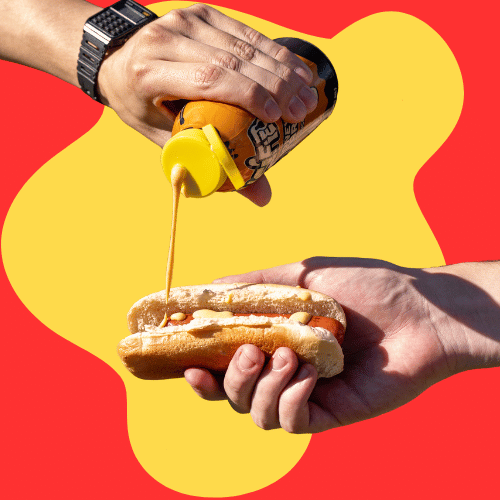 Hand pouring hot honey mustard onto a hot dog with a yellow and red background